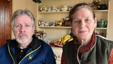 BBC David and Tracey sat next to each other; He is wearing a royal blue fleece and she is wearing an orange printed blouse with a khaki vest over it. They are sitting in front of a cabinet with teapots, cups and a fruit bowl.