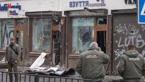 Anadolu via Getty Images Three people stand in front of a building with broken windows. Debris is strewn on the pavement in front of it.