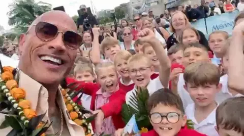 Disney A selfie-style image taken by The Rock – a man wearing glasses and smiling. Next to and behind him is a group of schoolchildren. They are cheering, smiling and shouting. They are wearing red and white school uniforms.