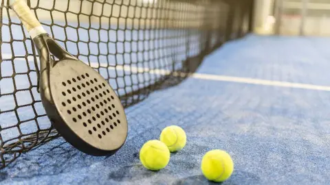 A black padel racquet leaning against a net with three tennis balls beside it on a blue padel court 