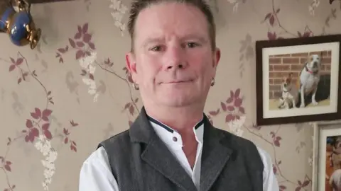 Lincolnshire Police A clean-shaven man with short brown hair and brown eyes is smiling at the camera in a living room decorated with a mauve and white leaf and flower design wallpaper. There is a framed picture of two dogs on the wall just above his left shoulder. He is smartly dressed in a grey sleeveless jacket and white collarless shirt and has an earring in each ear. 