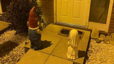 Helen Stark Image of a bride gnome and a gnome with glasses and a beard standing on a doorstep in the dark.