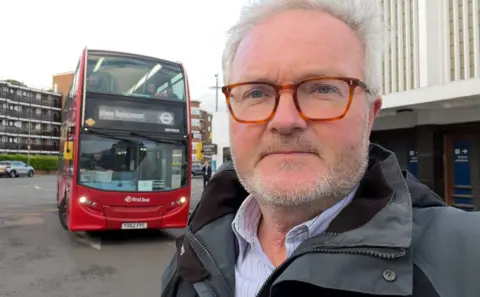 Peter Henley BBC South political editor Peter Henley wearing a grey jacket and blue stripped shirt stands in front of a red rail replacement bus