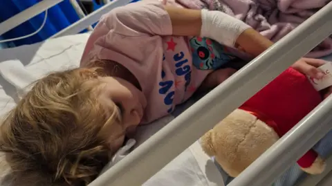 A young girl wearing Lilo and Stich pyjmas is laying in a hospital bed with a Winnie-the-Pooh teddy bear next to her.