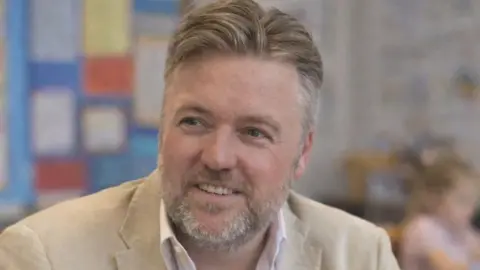 Anthony Holloway Anthony Holloway has short blonde hair and grey stubble. He is wearing a beige blazer over a white shirt. Out of focus behind him is a classroom.