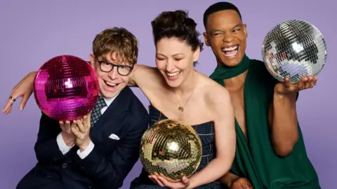 BBC Josh Widdicombe, Emma Willis and Johannes Radebe holding glitter balls against a purple background