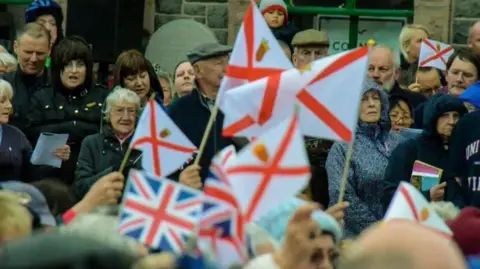 BBC A crowd of people standing outside holding Jersey and UK flags.