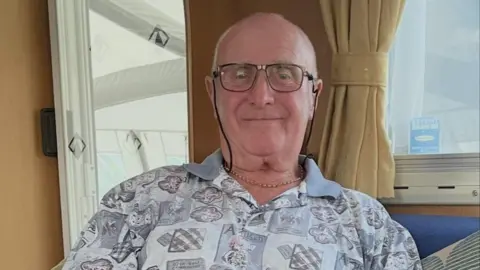 A man with cropped grey hair and glasses on a string is sitting on a caravan sofa with the awning visible through the windows. He is smiling at the camera and wearing a grey patterned shirt. 