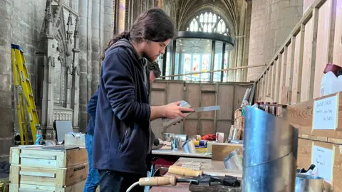 Organ builders work at a bench inside a cathedral, shaping metal components using tools laid out in front of them.