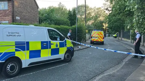 Merseyside Police officers and vehicles inside a cordon following an incident around the Tobruck Road area in Huyton on a sunny evening.