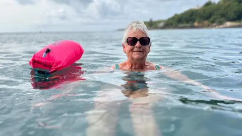 A lady submerged in water at Havelet Bay, she's wearing singlasses and has a pink buoy attached to her. She has short grey hair and is wearing a green swimsuit which you can see on her shoulders above the water. 