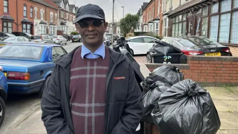 A man standing on a residential street next to rubbish bins that are overflowing with bin bags. He is wearing a black jacket with a red Prada logo, a burgundy jumper over a blue shirt, sunglasses and a black baseball cap. 