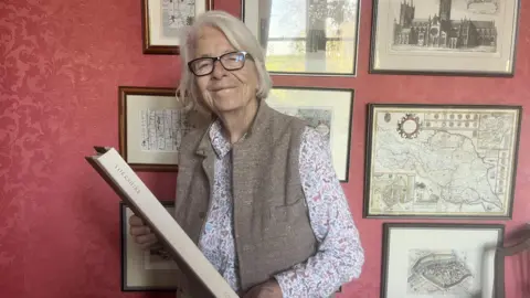 BBC A smiling woman with short grey hair wears black rimmed glasses, a gilet, and a floral shirt. She holds a large book and stands in front of a red-wallpapered wall, which is full of old, framed maps and photographs.