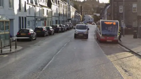 A street in a small town with old buildings and a bus and van coming towards us.