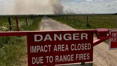 A red metal gate looking down a countryside track with large fields down either side and a large plume of smoke in the distance. Sign reads 'Danger impact area closed due to range fires'