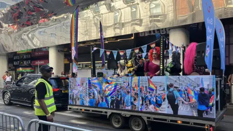 A trailer features several drag queens with microphones. One has huge red hair and a burgundy top. Another has a long pink ponytail and pink and black bodysuit. There are blue banners and rainbow banners across the top of the trailer, and large rainbow flags too