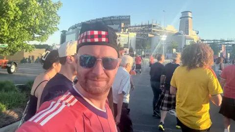 Man wears a Scotland top, hat and sunglasses, outside a sport stadium