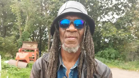 A man called Hubert Brown with dreadlocks wearing a hat and blue-tinted sunglasses looks at the camera.