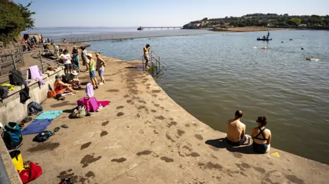 Swimmers sitting beside the marine lake in Clevedon, with various towels left around the outside of the pool, and the pier visible on the horizon in the background