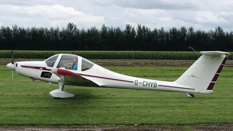 A Grob G109B motor glider from the side. It is white, with a round bubble cockpit, streamlined nose, slim tail and a large rudder. Inside is a man wearing a blue T-shirt. The glider is sitting on grass at an airfield. Crops and trees can be seen behind it.