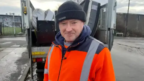 John Madden stands for the photo in front of a bin lorry. He's wearing a orange fleece and black beanie hat. 