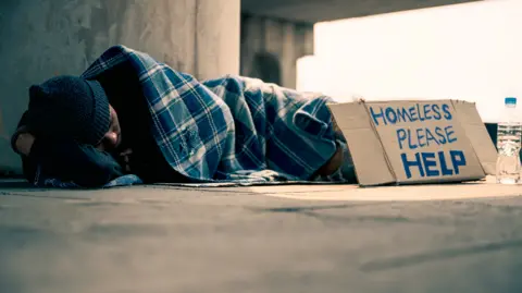 A young person sleeping on the streets, covered with a single blanket, with a 'homeless please help' sign propped against him.