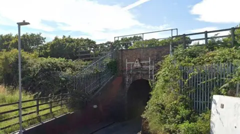 Google A narrow road passing underneath a small brick railway bridge surrounded by vegetation