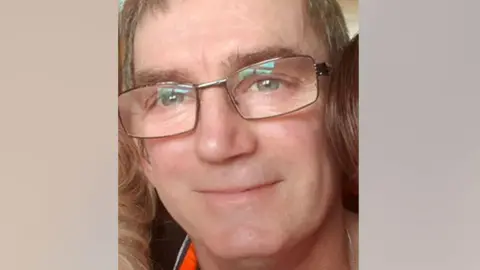 A man in his 50s with short hair and glasses smiles at the camera 