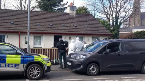 BBC Uniformed police officers and forensic investigators wearing white protective suits stand outside a house in Stream Street, Downpatrick. The house is a semi-detached or end-of-terrace bungalow. There are trees behind the houses and church with a tall spire in the distance. A liveried blue and yellow police car is parked outside the house and other vehicles are parked in the street.