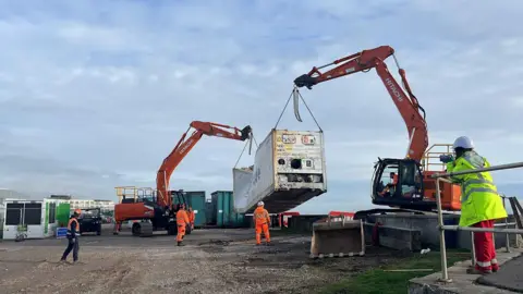 George Carden/BBC Two excavators hold up the container on chains in a nearby car park.