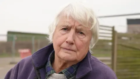 Shaun Whitmore/BBC Bryony Nierop-Reading, a woman with short grey  She is wearing a blue and green checked shirt with a cream jumper. She is also wearing a purple fleece and is looking into the camera. 