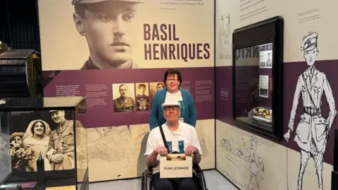 BBPR Leonard Symonds, 88, wearing a white t-shirt posing for a photo at the Bovington Tank Museum. He is wearing glasses and is smiling at the camera.