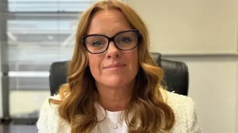 Claire Christian, who has auburn hair and is wearing large dark rimmed glasses and a white top with a cream jacket, sitting in a black chair in an office.
