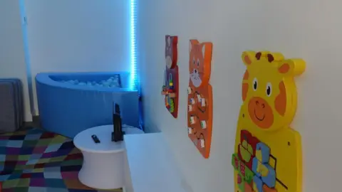 A room with brightly-coloured wall-mounted puzzle games, a ball pool and fluorescent blue light in the background.