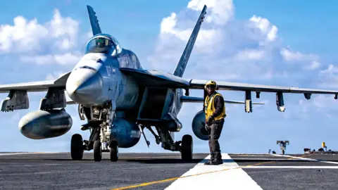 In this handout photo provided by the U.S. Navy, A U.S. Sailor signals to an F/A-18E Super Hornet aircraft, attached to Strike Fighter Squadron 87, on the flight deck of the world's largest aircraft carrier, USS Gerald R. Ford (CVN 78), while operating in support of Operation Epic Fury on March 2, 2026 in the Mediterranean Sea.