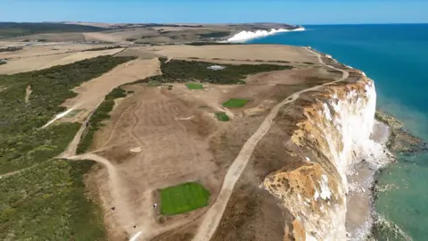 A white cliff face at Seaford Head, where the path and grassland on top appears dry and brown.