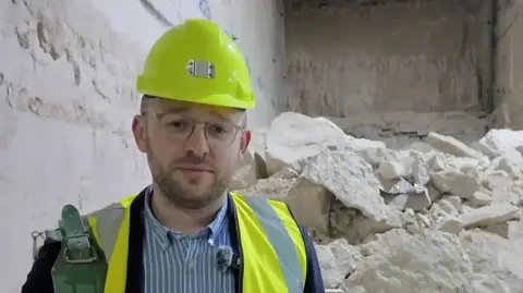 Lloyd Hatton is wearing a yellow hi-vis vest and hardhat, with a blue and white stripy shirt and navy blazer. Behind him is a pile of quarried white stone.