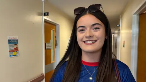 A teenage girl with long brown hair wearing a blie and red t shirt, a pendant in the shape of the sun and a sunglasses on her head. She is smiling and look at the camera.