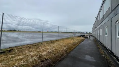 Glyn Matthews A wet path runs between a grey portable office building and a high wire fence. Beyond the fence lies a wide, empty runway under an overcast grey sky. Scrubby grass and picnic benches line the edge of the path.