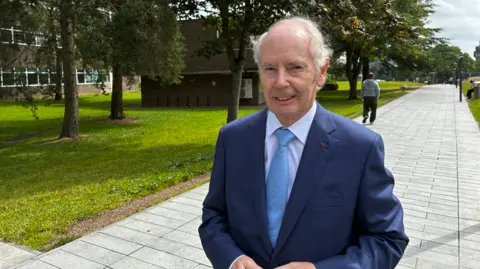 A man in a suit stands on a paved walkway beside trees and grass, with buildings in the background.