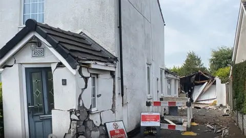 BBC A white rendered house with a porch with a blue door that has massive cracks on it and is reduced to rubble in the right corner. To the side is a driveway with a broken garage and red and white signs warning not to smoke