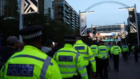 Met police officers, wearing bright yellow jackets, walk in front of Wembley stadium