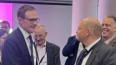 A man with glasses and a suit smiles as he shakes hands with another man in a suit at the tory conference. They are surrounded by other men attending the conference.