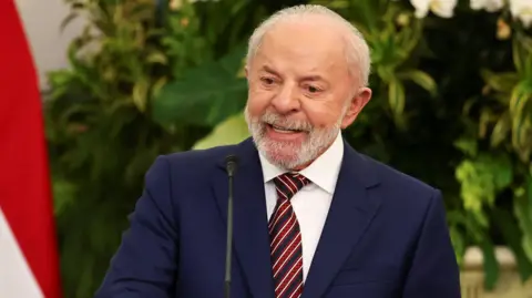 Reuters Lula is wearing a fitted navy suit with a white shirt on underneath. He is wearing a funky red, white and blue striped tie and is standing behind a lectern in mid speech. He has a grey beard and grey hair. 