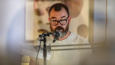 Stuart Whipps in a white t-shirt speaking into a microphone, with an indistinct projection behind him