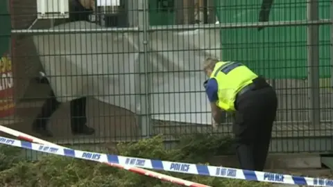 A police officer leans over next to a metal fence which is behind police tape