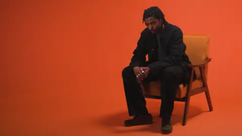 A man dressed in black sits on a wooden chair against an orange background