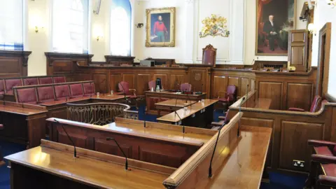 BBC Guernsey States' debating chamber. A wood-panelled room with oil paintings, multiple red leather-upholstered seats and tables, plus several large windows.