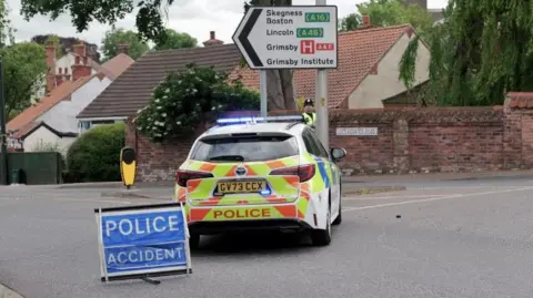 Grimsby News & Pictures Agency A police car blocks a road close to the scene of an accident. A brick wall and road sign are visible in the background alongside a row of houses. A blue sign which reads "Police Accident" has also been place in the road.