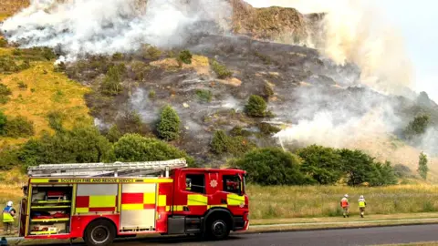 Fire trucks parked at foot of hill which is on fire